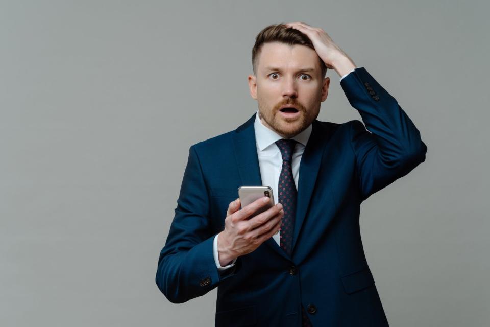 Shocked investor with open mouth, hand up, and smartphone in one hand.