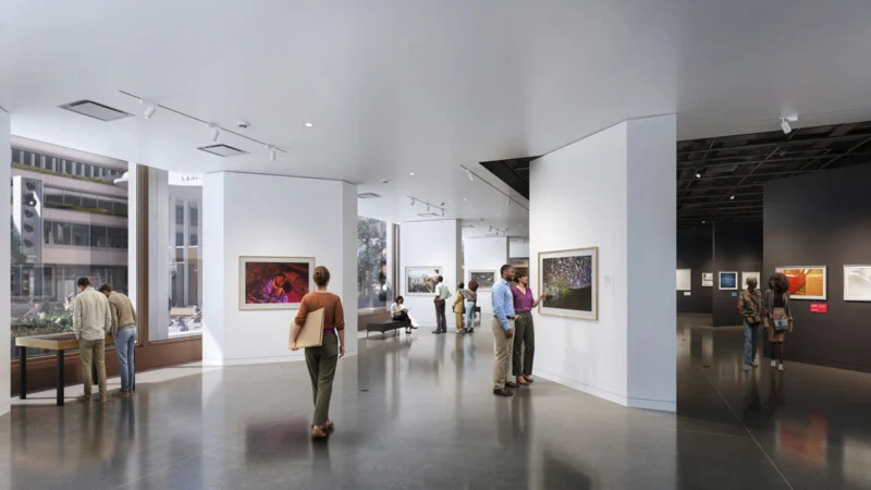 People watch movies in a bright, modern art gallery with large windows, white walls, paintings and sculptures. Some visitors stand and watch, while others chat or sit on benches.