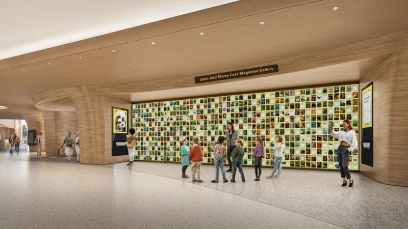 A group of people, including children, are looking at a large storage wall filled with colorful magazine covers in a wide, modern hallway. "Joan and Steve Case Magazine Gallery.