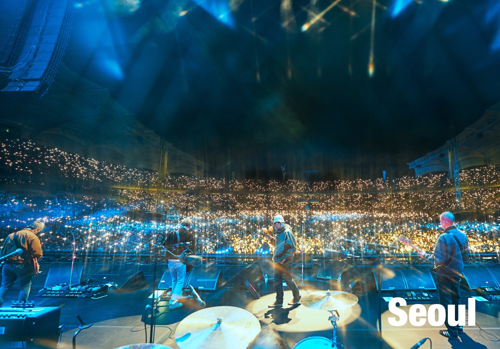 A wide, high-color shot shows the band playing on a big stage in Seoul, facing an arena filled with thousands of bright string lights.