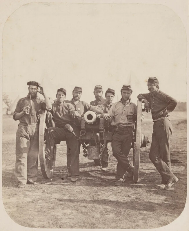 Seven Civil War soldiers in uniform stand around a large cannon in a grassy field, with several tents visible in the background. Others lean in a random manner against cannon wheels.