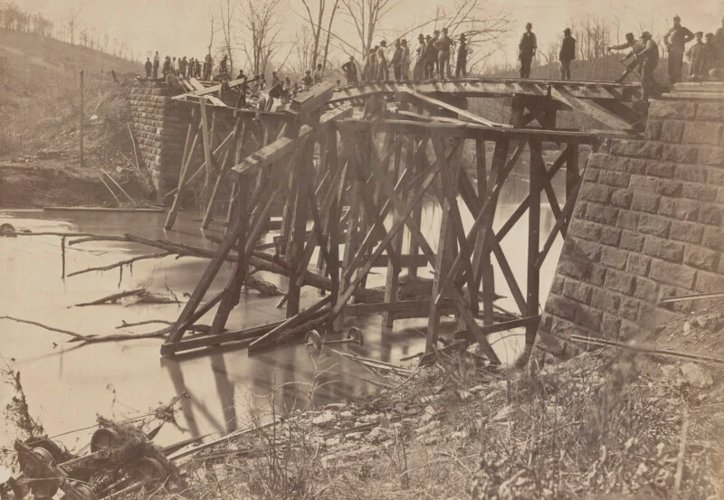 A group of people stand on and around a dilapidated wooden bridge over a river, with fallen parts and debris in the water, surrounded by bare trees and rocky cliffs.