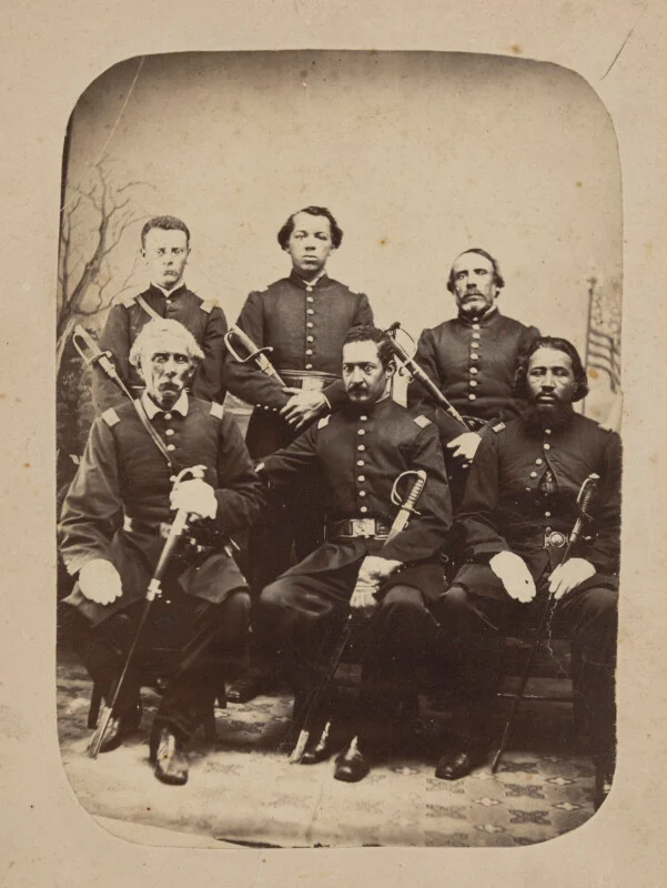 Six men in Civil War-era military uniforms form a formal portrait. Three are sitting in front holding swords, while three are standing behind them. The American flag is slightly visible in the background.