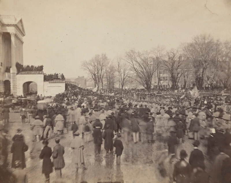 A large crowd gathers outside the large pillared building on a winter day, many wearing coats and hats. Leafless trees and more buildings are visible in the background.