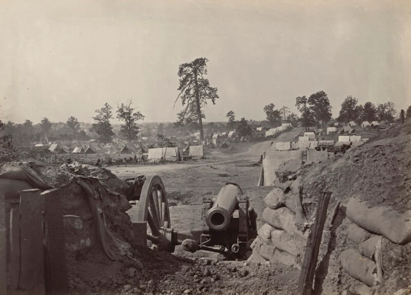 A black-and-white photo shows a Civil War-era military camp with tents spread across a field. In front, there are large cannons placed behind sandbags and earthworks. Trees surround the camp in the distance.