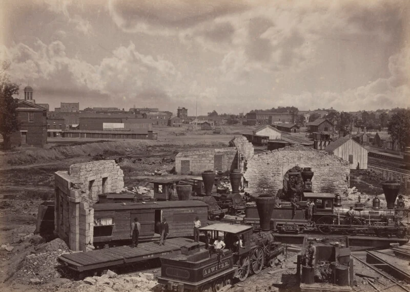 Black and white photograph of a 19th century scene with ruined stone buildings, many steam trains, and people standing by and around them; city ​​buildings and cloudy sky in the background.