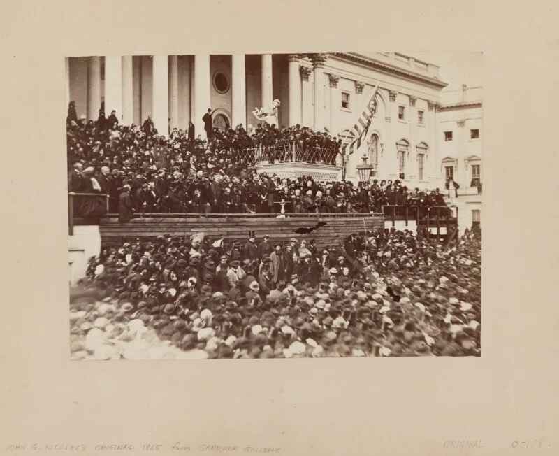 A large crowd gathers outside the government building for the event, with people standing on the steps and balconies. An American flag hangs nearby, and a speaker addresses the audience from the stage.