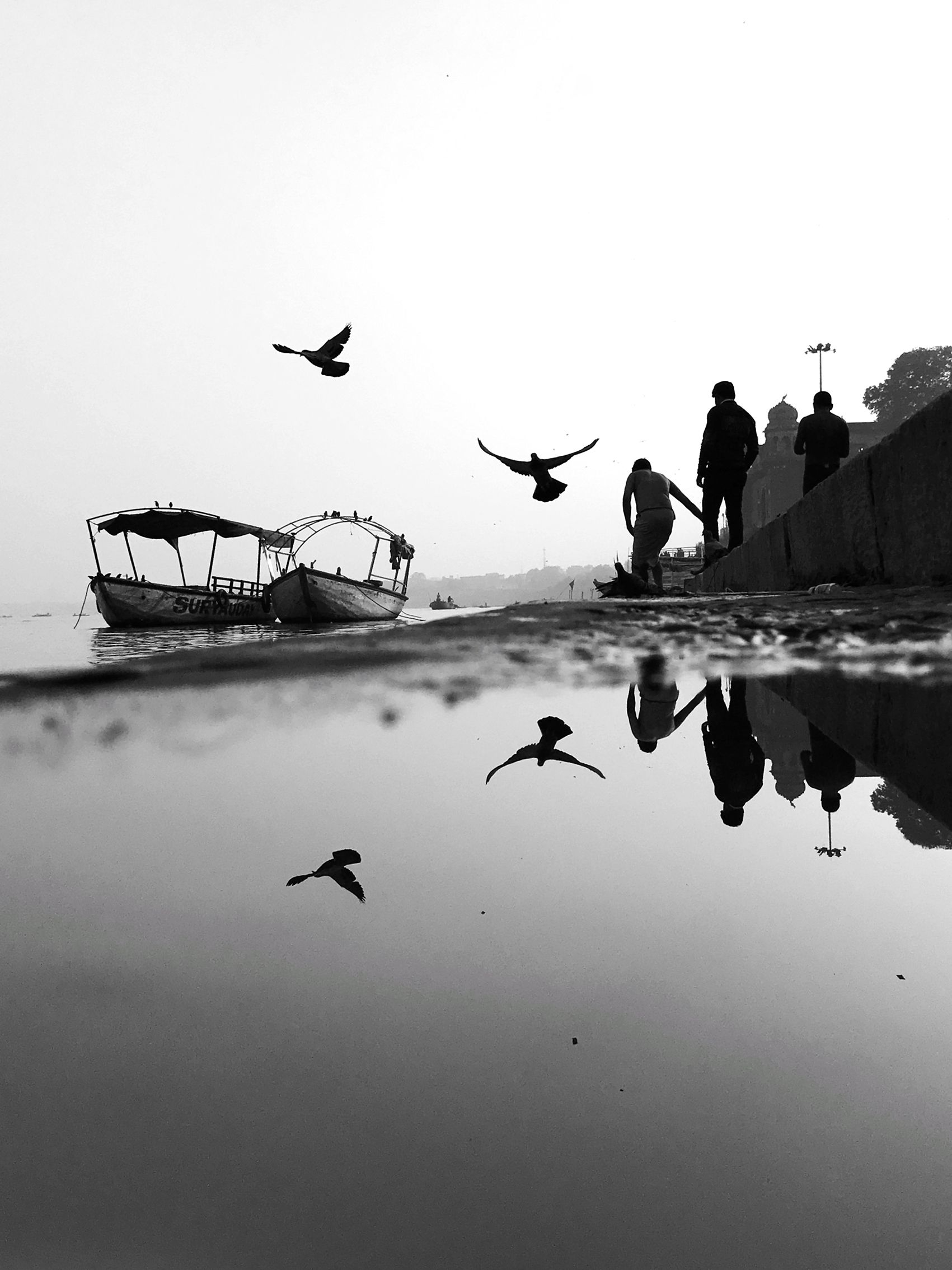 Black and white image of a riverside scene with boats, two pedestrians and birds flying overhead. Their expressions are reflected in the water hole