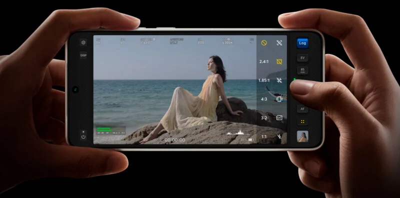 A man holding a smartphone above, taking a woman in a yellow dress sitting on the rocks of the sea, with the camera settings on the screen.
