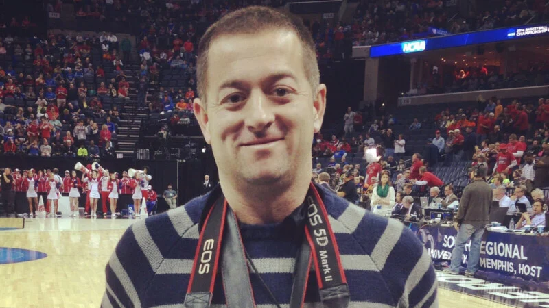 A man wearing a colorful sweater and a camera around his neck is standing on a basketball court, with spectators and players visible behind him.