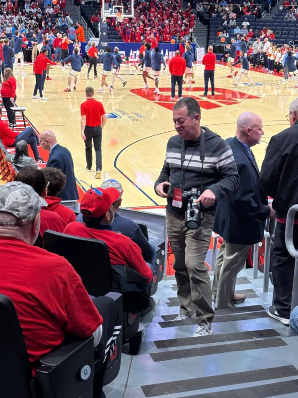 A photographer stands on the steps of the stadium with a camera, talking to a seated fan. Red-shirted fans fill the stands, and basketball players warm up on the court behind them.