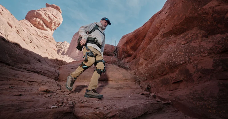 A man wearing hiking clothes and a backpack walks in a rocky desert canyon, surrounded by red sandstone cliffs under a blue sky.