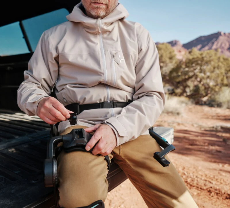 A man in a light jacket and brown pants sits on the bed of a truck outside, adjusting a black wall on his leg in a rocky, desert-like landscape.