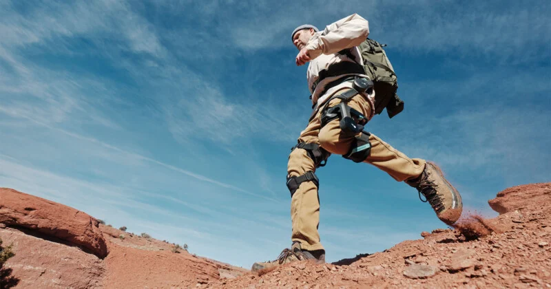 A man in outerwear and a rucksack walks up a rocky hill under a clear blue sky, concentrating on his feet as he walks with the dirt from his boots.