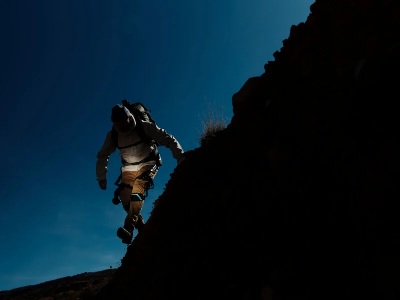 A man in outerwear and a rucksack climbs a steep rocky slope against a clear blue sky, slightly lit by sunlight.