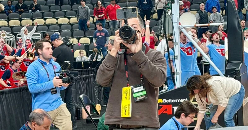 The photographer pans the camera to a spectator on a basketball court, with a sports crowd and band singing in the background. Other media workers and event workers are also seen surrounding him.