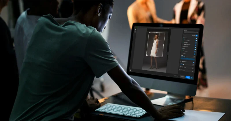 A person uses a computer to edit a photo of a woman in a white dress; some people and studio lighting equipment are visible in the background blur.