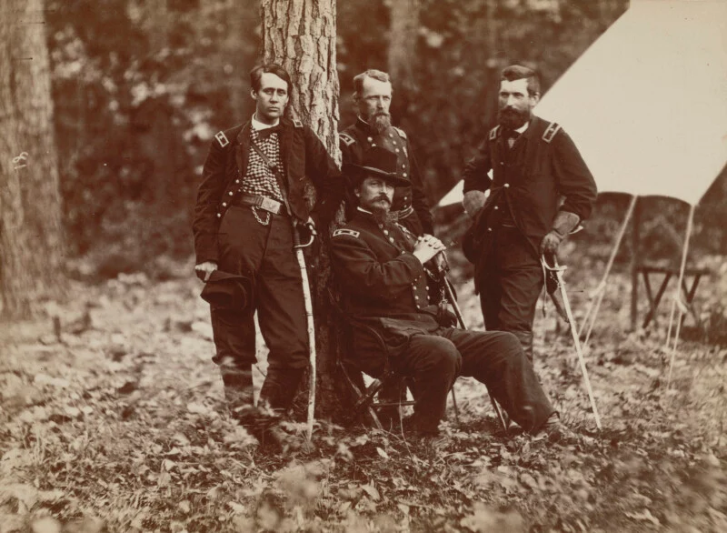 Four men in 19th century military uniforms stand in a wooded camp. Three are standing next to the tree while one is sitting in front, near the tent. Fallen leaves cover the ground.