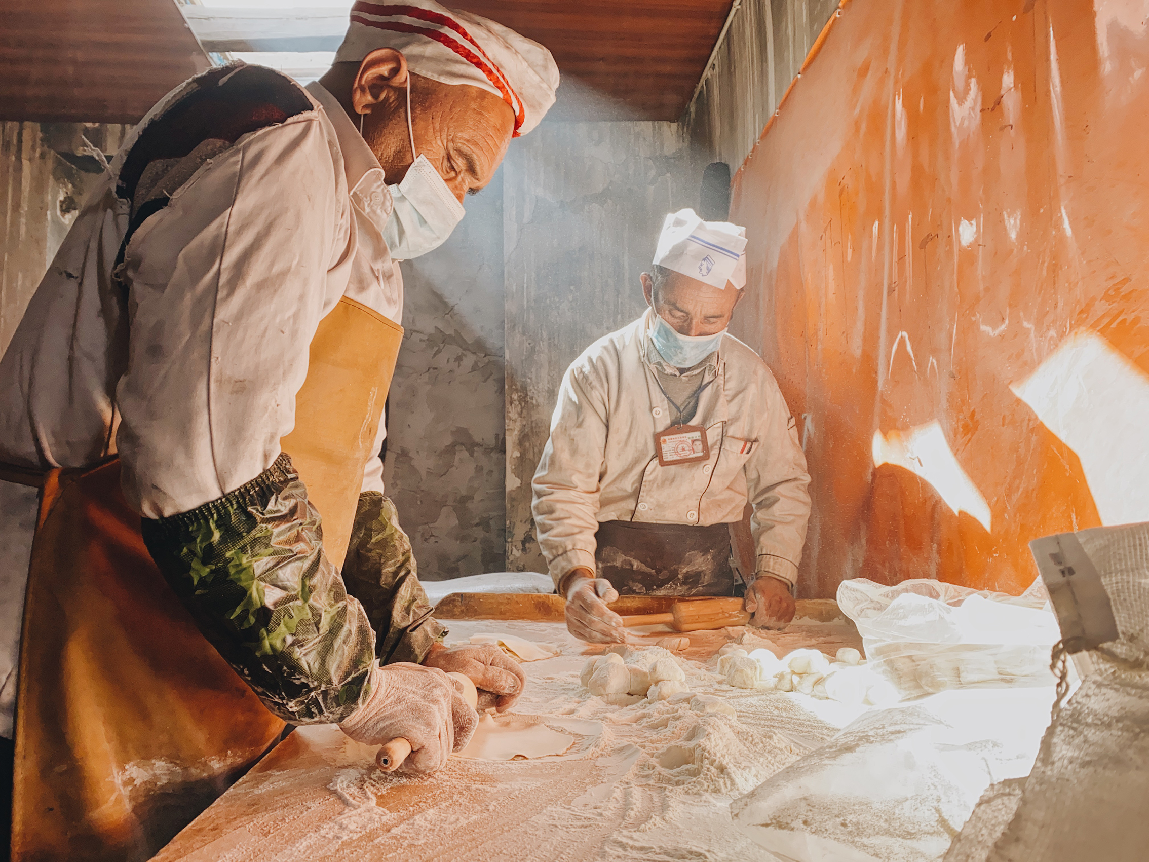 Two chefs in aprons and masks are hard at work in a sunlit kitchen, rolling and forming dough on a floured table.