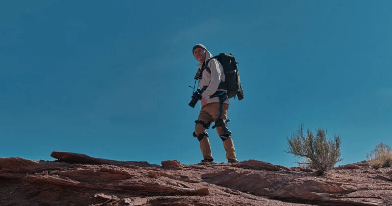 A man wearing outerwear and a backpack is standing on a rocky outcrop under a clear blue sky, holding a camera and looking into the distance. Vague desert vegetation soon appears.