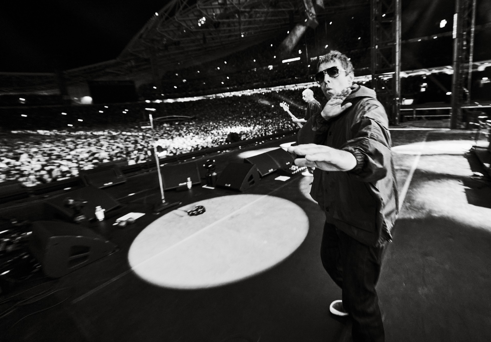 Liam Gallagher leans into the camera with his arms outstretched during a performance in a large, crowded arena, captured in stark black and white.