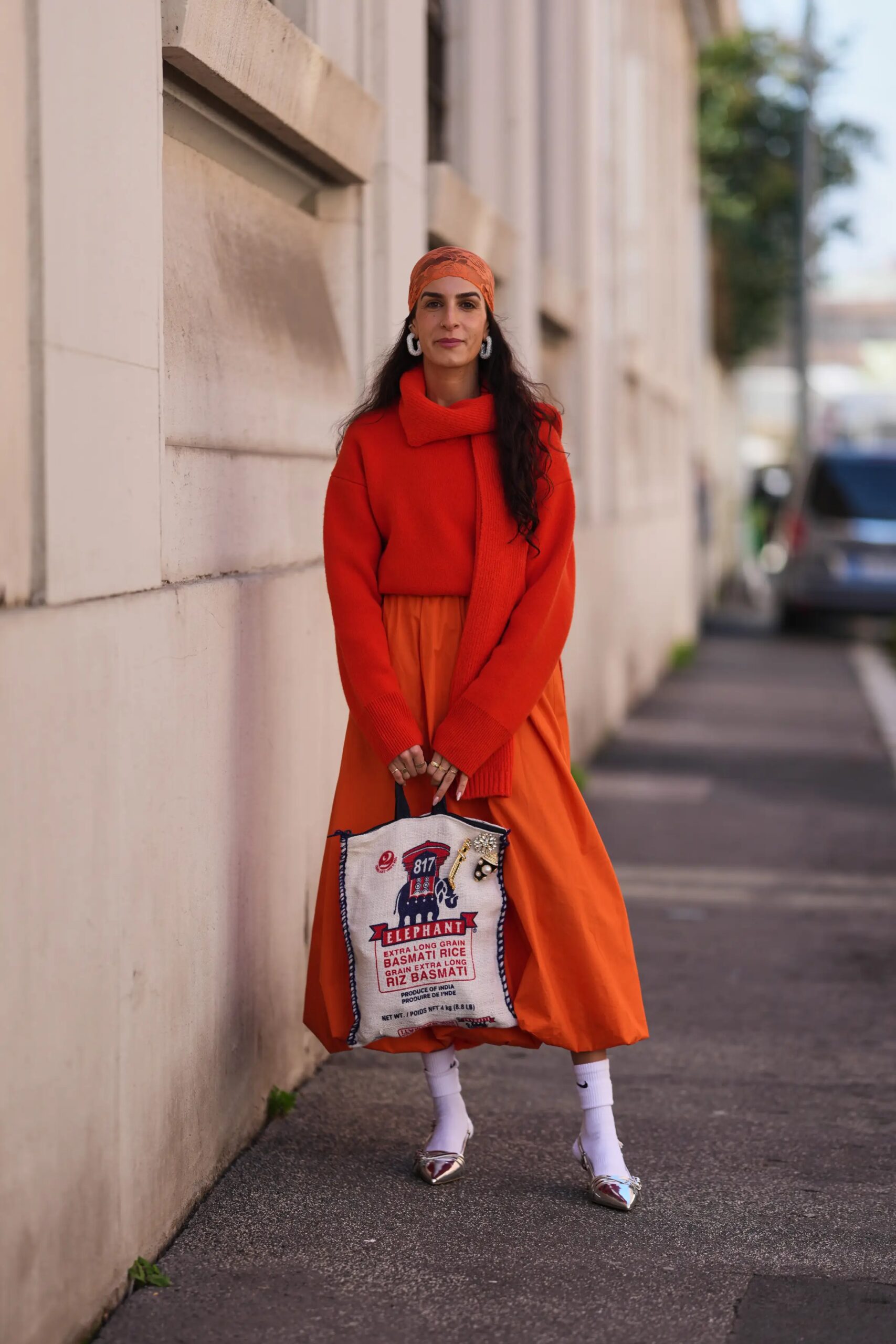 A woman wearing an orange scarf, a bright red knitted sweater, an orange satin midi dress, and carrying a bag shaped like a rice bag.