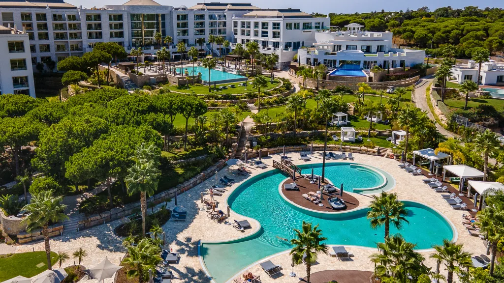 A resort pool with a central deck, lounge chairs, cabanas, palm trees, and a modern hotel complex in a bright tropical setting.