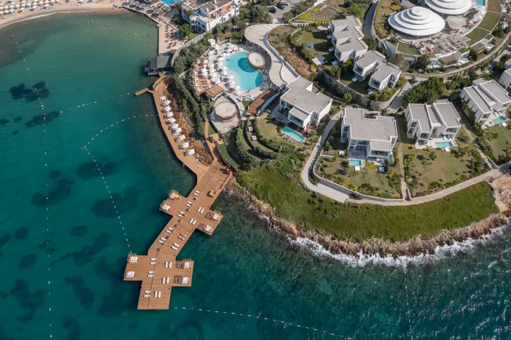 A beach resort with white buildings, private pools, a wooden pier with loungers, a lap pool, and rocky beaches close to the ocean.