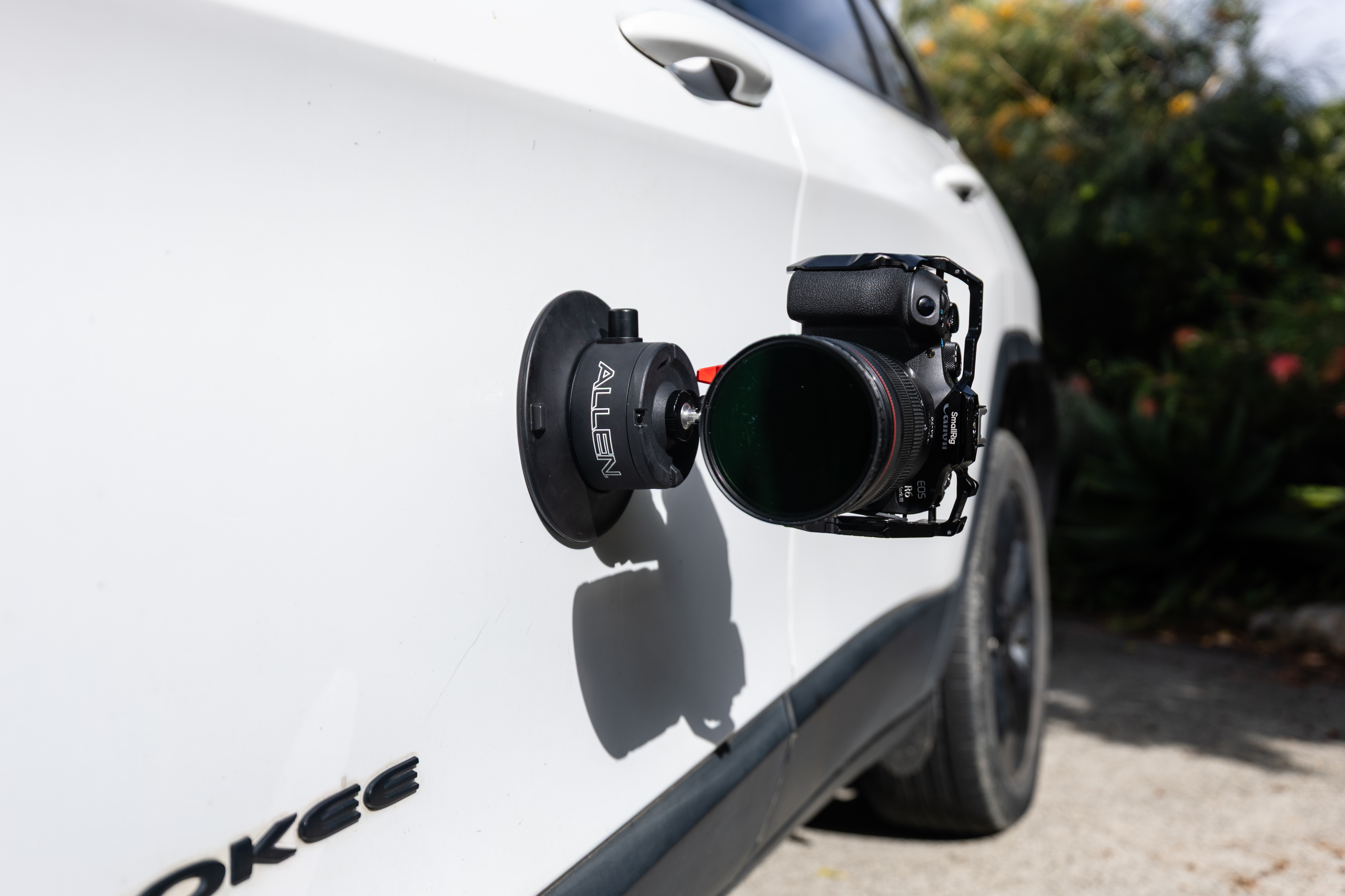 The Allen Smart Suction Snap Camera Mount is mounted high on the side of a white Jeep, securely holding a Canon mirrorless camera with a professional lens.