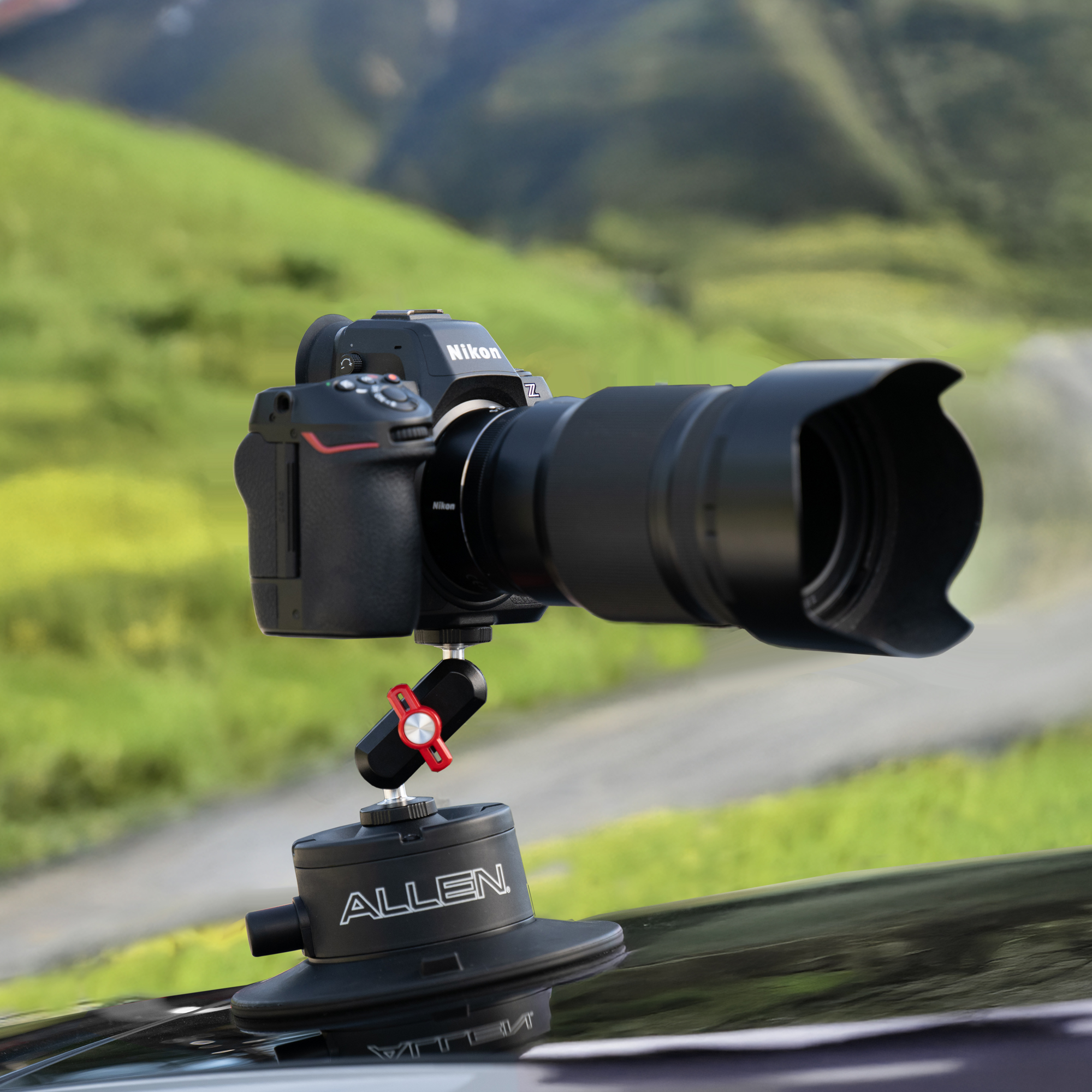The Allen Smart Suction Snap Camera Mount is attached to the hood of a black car, supporting a Nikon mirrorless camera with a large lens against a backdrop of green hills.