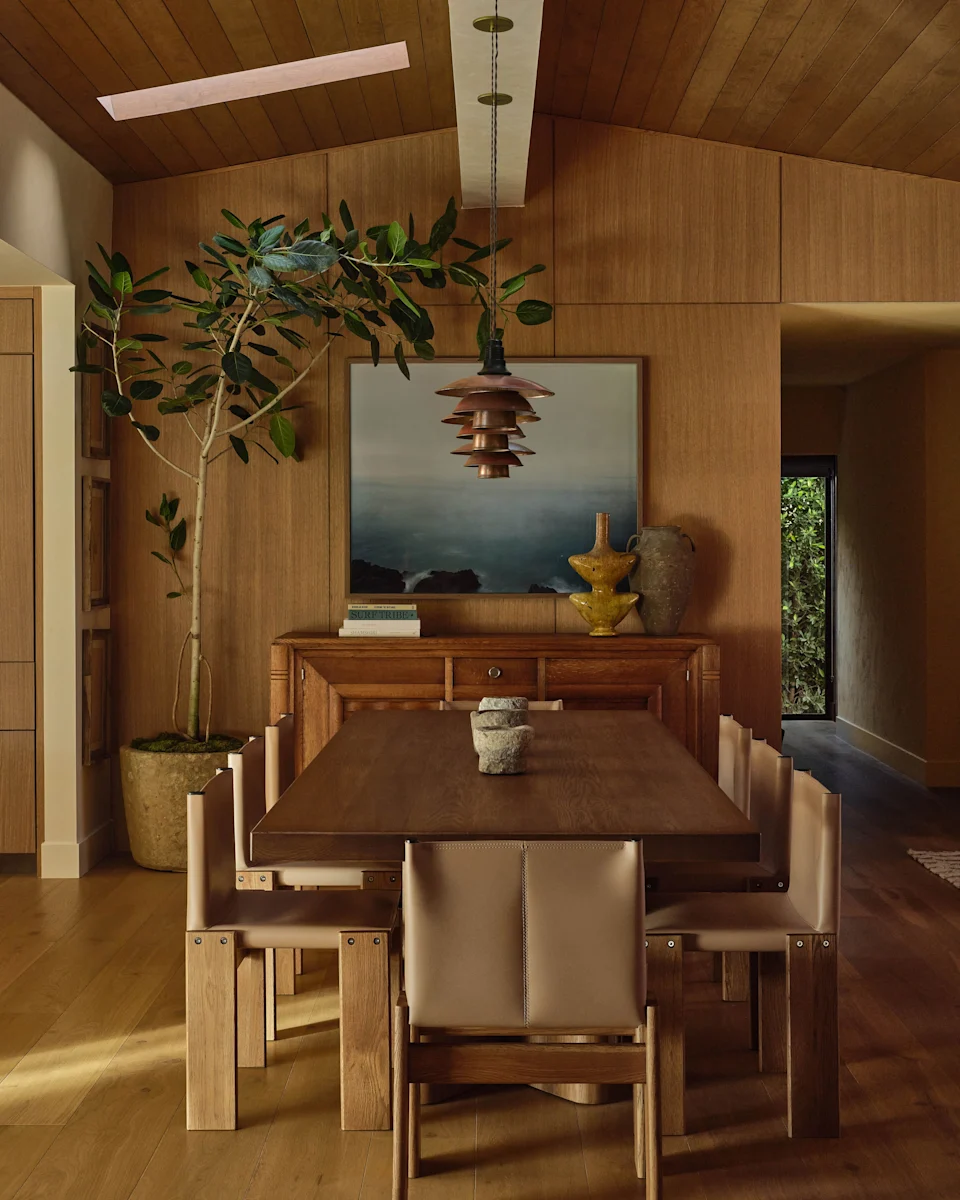 In the open heart of the house, a formal dining table in the style of Pierre Jeanneret sits under a coffered ceiling, surrounded by chairs by Afra and Tobia Scarpa Monk. An evocative photograph of the Pacific Ocean by Rennie Solis hangs above a Charles Dudouyt French oak 1940s buffet by Lone Fox. Solis designed a series of antique bronze paintings by Poul Henningsen for Louis Poulsen. Ficus Audrey from Inner Gardens is placed in a limestone base by Olive Ateliers.