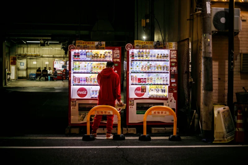 A man dressed in red stands at night in front of two bright vending machines full of drinks, on a dark street with bars in front of the machines.