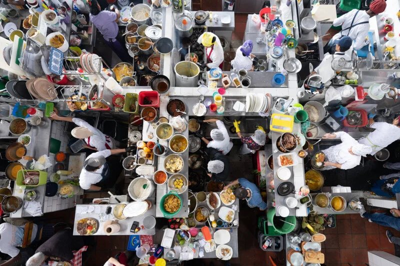 A bustling scene of chefs working at separate kitchen stations, filled with pots, pans, plates and ingredients, creating a vibrant and chaotic cooking environment.