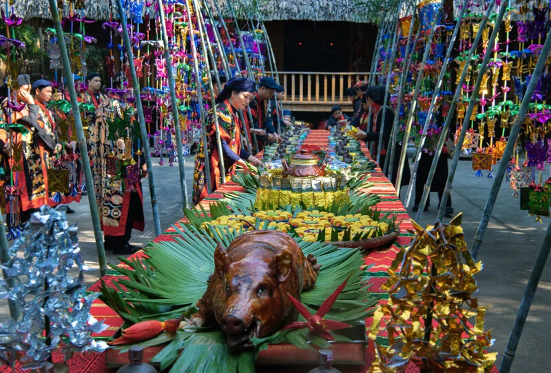 A festive, colorful table is set outside with a roast pig as the centerpiece, surrounded by traditional decorations, tropical leaves and food dishes. People in strong traditional clothes stand on both sides.