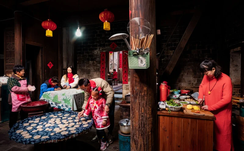 The family gathers in a common kitchen with warm lights, preparing food together. Children and adults work at tables with dough and utensils, while red lights and festive decorations hang around the rustic room.