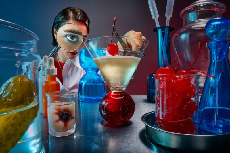 A woman in a white coat and holding tweezers puts a cherry in a martini glass. Large glass jars and beakers containing cherries, eyeballs, pickles and blue liquids surround him on the laboratory table.