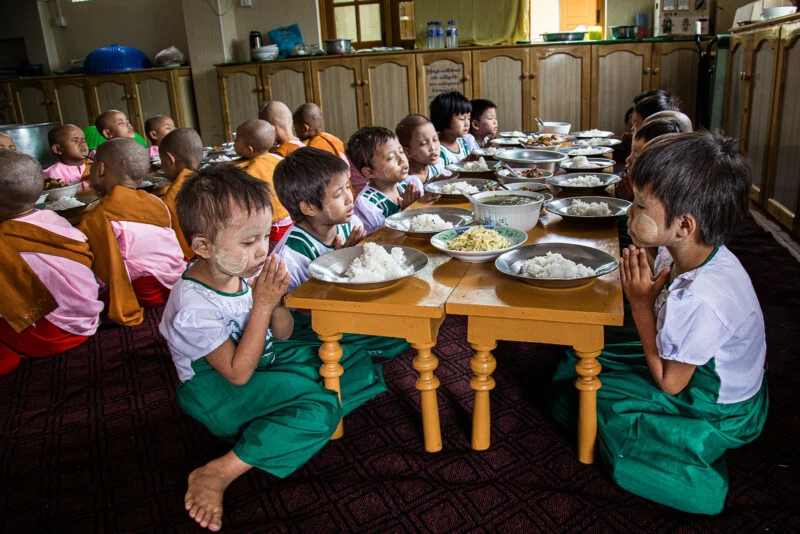 Small children in traditional face painting sit cross-legged in groups around low tables, hands folded in prayer, with bowls of rice and food in front of them, in the communal dining hall.