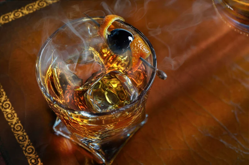 A cocktail smoked in a glass with large ice cubes, garnished with an orange twist and a dark cherry to choose from. Wisps of smoke rise from the drink, which rests on the decorated leather.