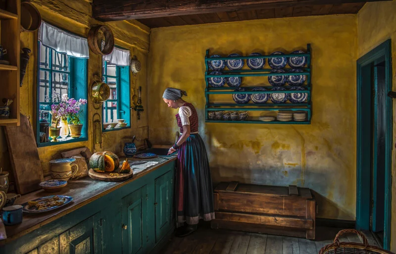 A woman in traditional clothes stands by a window in an old kitchen, with yellow walls, blue cupboards, flowers and shelves with blue and white dishes. Sunlight streams through the window.