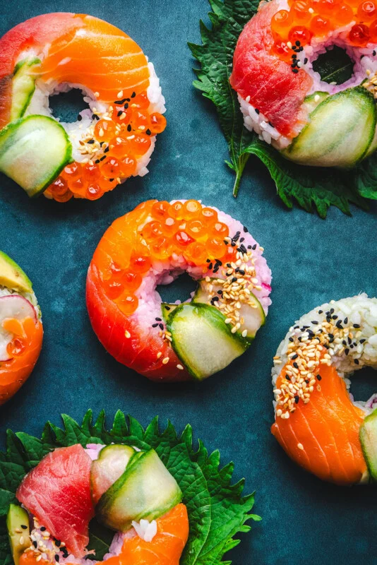 Colorful donuts topped with slices of salmon, tuna, cucumber, sesame seeds and orange fish roe, arranged on dark blue with green leaves underneath some of the sushi pieces.
