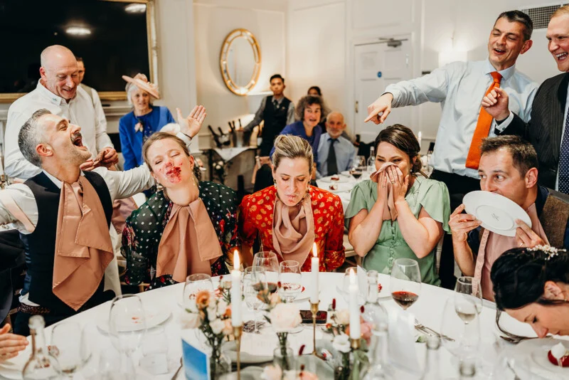 A group of well-dressed people sit around a table at a festive event. Some have cream or dessert on their faces and are laughing, while others cover their faces in amusement. The atmosphere is fun and festive.