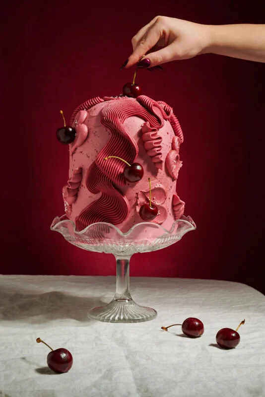 A tall pink cake decorated with cherries sits on a glass stand against a dark red background. A hand places a cherry on top, and more cherries are scattered on the white tablecloth below.
