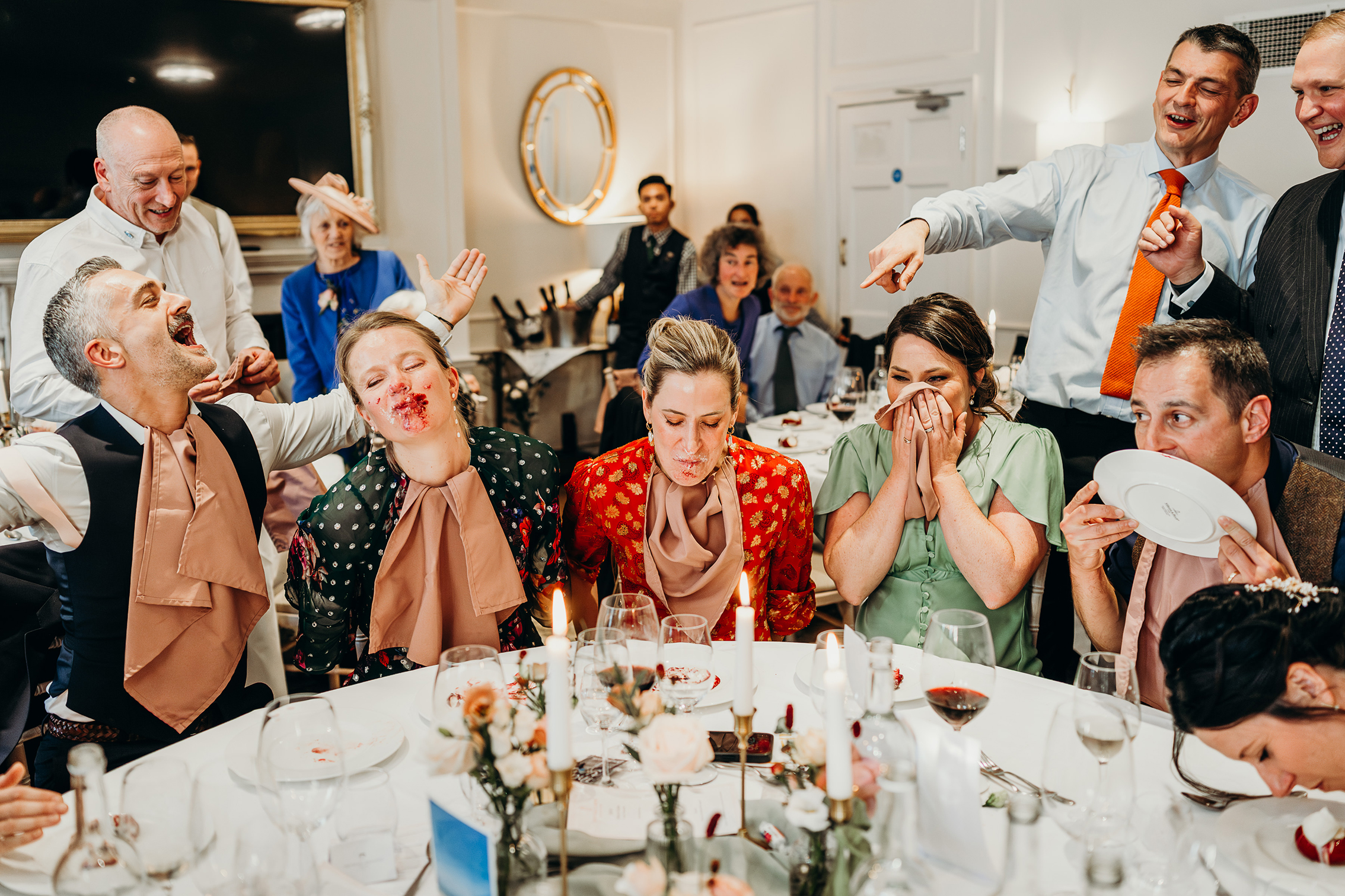 A fun scene at a dining table shows people laughing and playing a game with blindfolds and bibs. Some have food on their faces