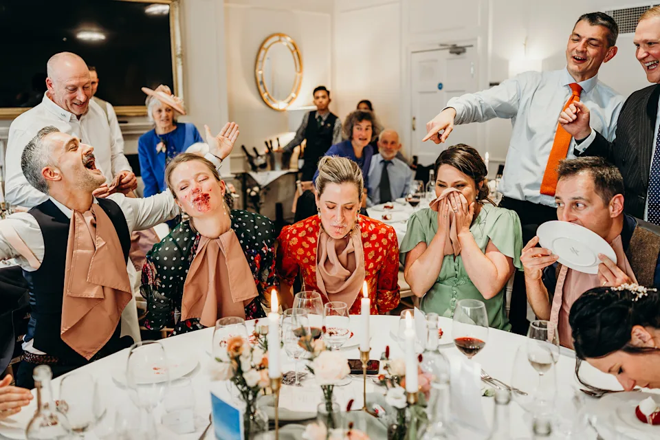 A fun scene at a dining table shows people laughing and playing a game with blindfolds and bibs. Some have food on their faces