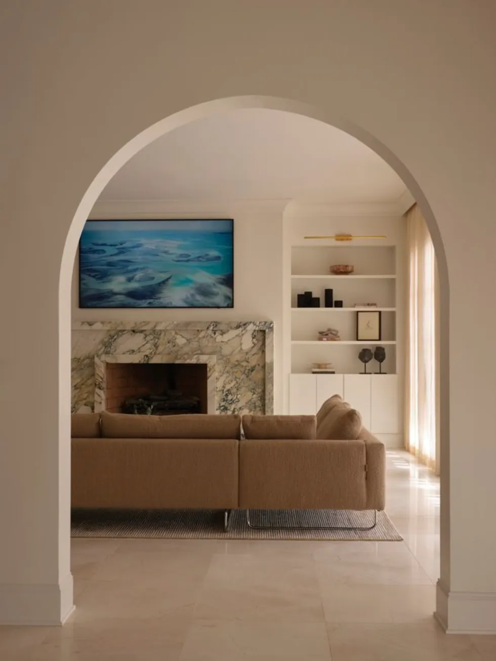 A cozy living room with arched doors, a brown sofa, a marble fireplace, and a wall-mounted TV.