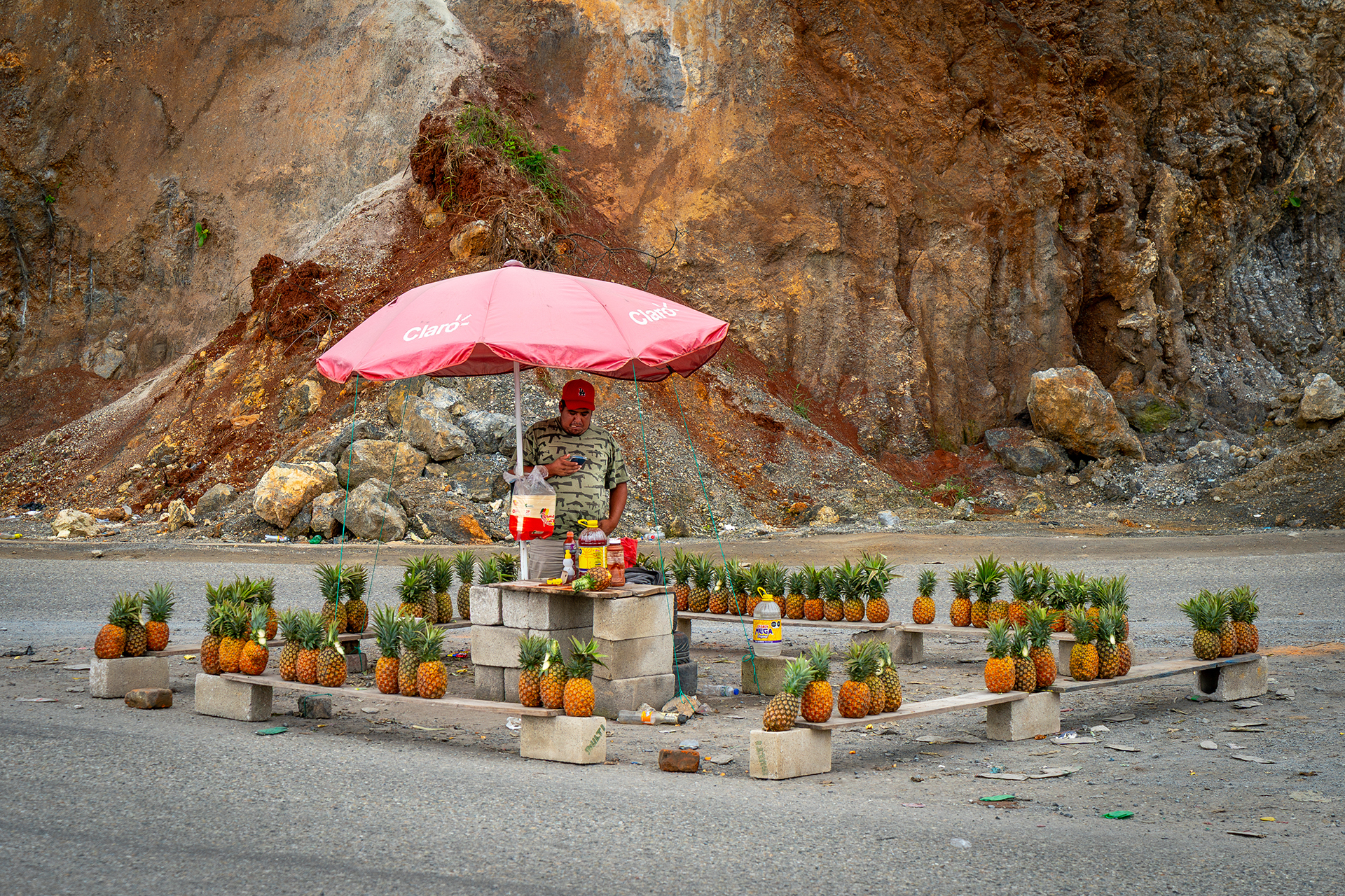 A pineapple chair by the road and a man under a pink umbrella. The bench is made of cinder blocks, and the pineapples are arranged neatly in the rocky area